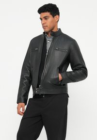 Juodas odinis švarkas su aukštu apykakle, užtraukiamomis kišenėmis ir raukšlėmis. Lygus paviršius su struktūruotu sijonu, dėvimas ant languoto marškinėlių.