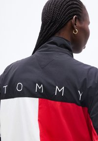 Άτομο με πλεξούδες που φοράει μαύρο, λευκό και κόκκινο μπουφάν με το κέντημα "TOMMY" στην πλάτη και χρυσό κρίκο στο αυτί.