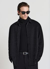 Camicia nera con bottoni e una finitura leggermente testurizzata, indossata sopra un dolcevita nero aderente. Il look è completato da occhiali da sole neri.