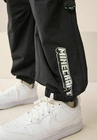 Fekete jogging nadrág rugalmas mandzsettákkal, az oldalán fehér "Minecraft" logóval, fehér sportcipővel párosítva. Sima textúra.