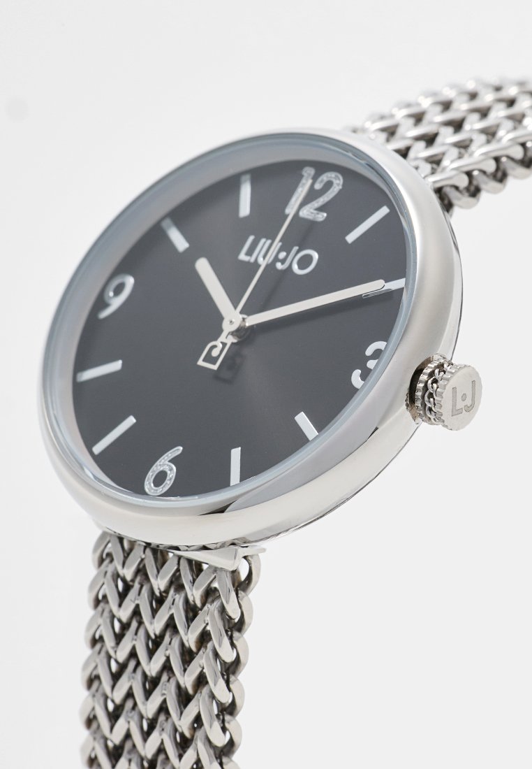 Zegarek na srebrnej siateczkowej bransolecie z okrągłą czarną tarczą, białymi znacznikami godzin i wyraźnym logo "LIU JO". Posiada srebrną koronę z grawerem "LJ".