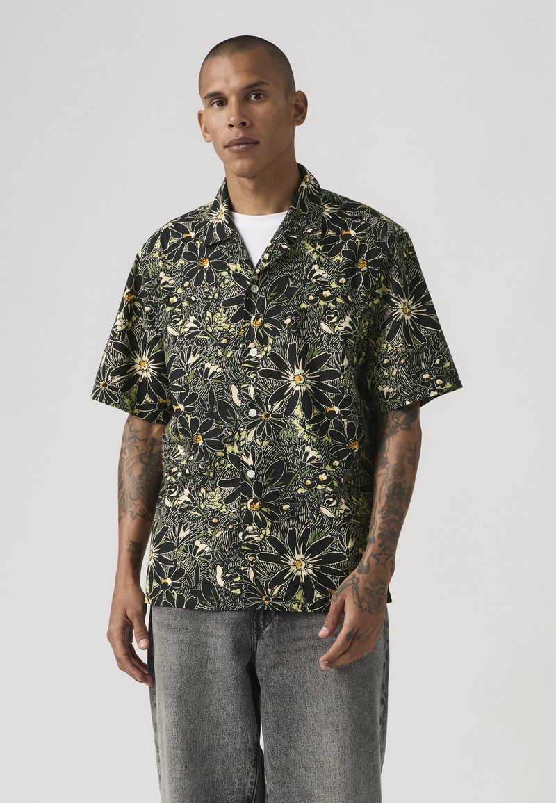 Uomo con tatuaggi che indossa una camicia nera a maniche corte con bottoni, decorata con grandi motivi floreali gialli e bianchi, e jeans grigi.