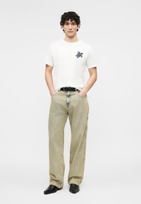 Joven con cabello rizado que lleva una camiseta blanca con un dibujo de tortuga, pantalones anchos beige a rayas, cinturón negro y zapatos negros puntiagudos.