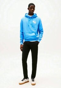Young man wearing bright blue hoodie, black pants, and white sneakers standing against plain white background.