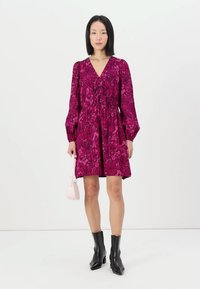 Robe à manches courtes, fuchsia, avec un motif floral, taille cintrée et décolleté en V ; associée à des bottines noires et un petit sac à main rose.