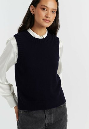 Donna che indossa un maglione smanicato blu navy sopra una camicia bianca con bottoni e jeans grigio scuro su uno sfondo chiaro e semplice.