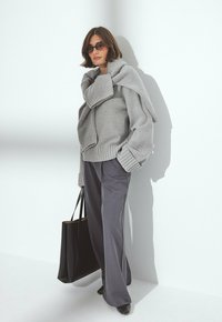 Vrouw in een oversized grijze trui met een bijpassende gedrapeerde trui, brede grijze broek, zonnenbril, en houdt een grote zwarte schoudertas vast.