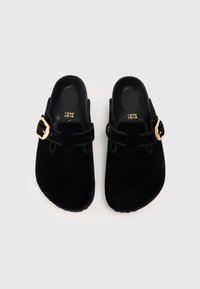 Sabots en suede noir avec bout rond, sangle réglable et détail de boucle dorée. Texture douce avec un intérieur rembourré. Taille 37.