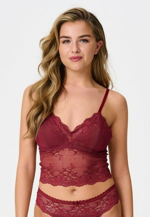 Femme aux longs cheveux ondulés portant un ensemble de lingerie en dentelle rouge composé d'un bralette et d'un bas assorti sur un fond uni.