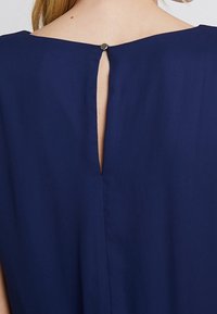 Robe bleu marine avec une ouverture en trou de serrure dans le dos, fermée par un bouton en métal, présentant une texture lisse et un design drapé.