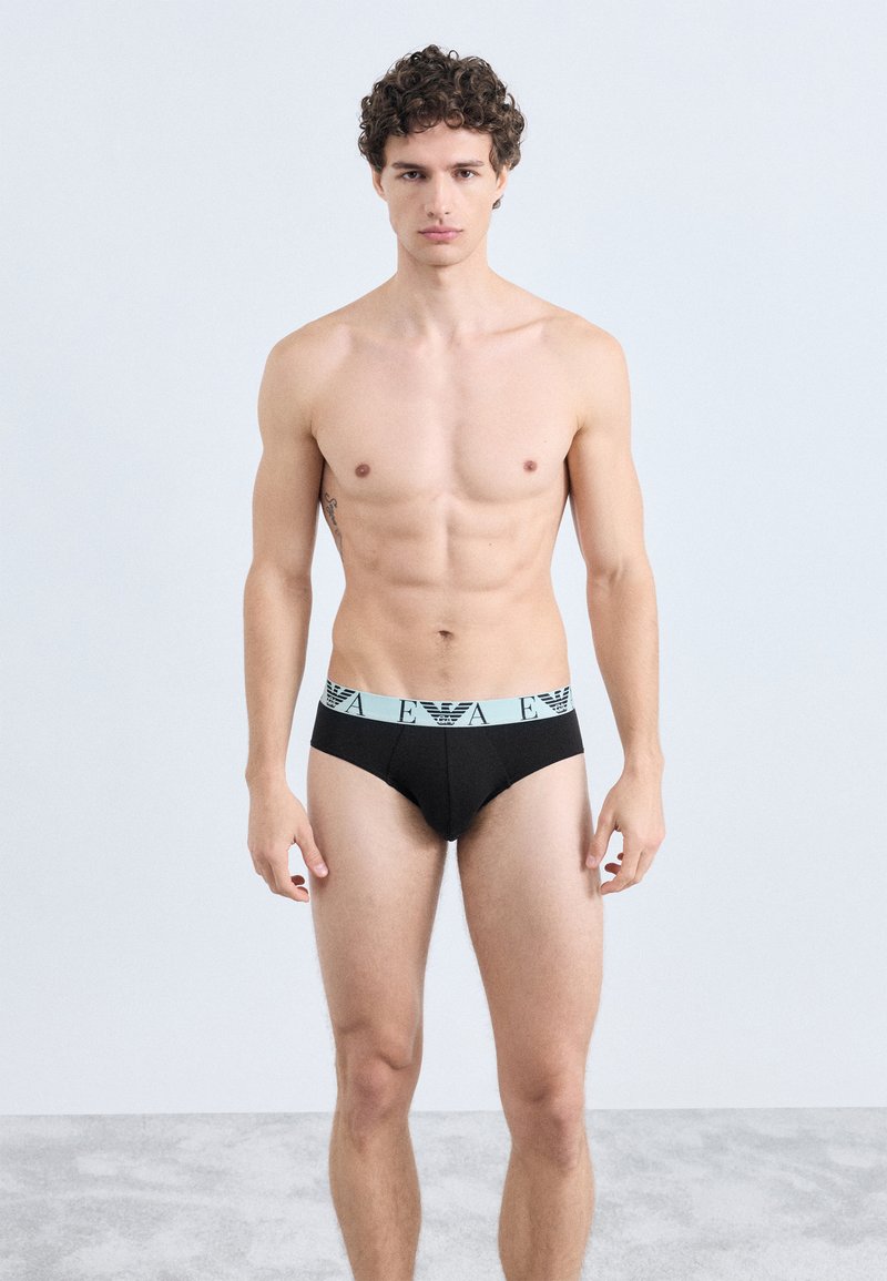 Cuecas pretas com cós elástico azul claro com um logótipo. Modelo em pé, mostrando o ajuste e o design contra um fundo simples.