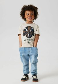 Lysbeige t-shirt med en brun dinosaur grafik, parret med lyseblå cargo bukser med snøre i taljen og sorte sneakers.
