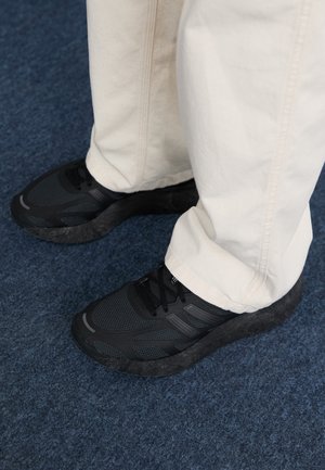 Personne portant des baskets noires et un pantalon écru debout sur un tapis bleu.