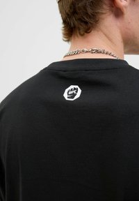 Sort kortærmet t-shirt lavet af bomuld, med et hvidt UFC-logo i en oktagonform på ryggen nær halsudskæringen.