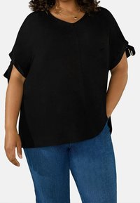 Black V-neck top with short, rolled sleeves and side slits, styled with contrasting accents. Paired with blue denim jeans.