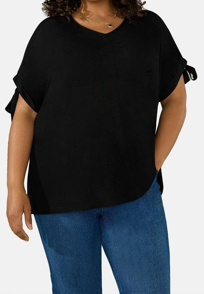 Black V-neck top with short, rolled sleeves and side slits, styled with contrasting accents. Paired with blue denim jeans.