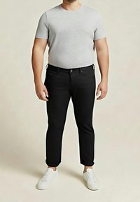 Homme portant un t-shirt gris clair à manches courtes, un pantalon noir et des baskets blanches, debout devant un fond uni.
