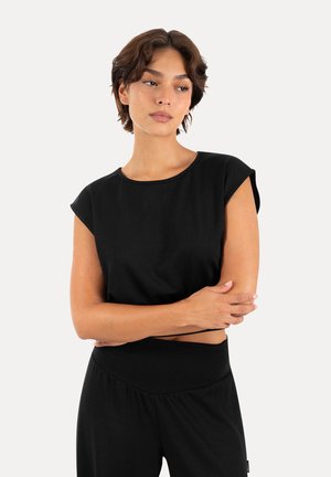Jeune femme aux cheveux bruns courts portant un haut noir sans manches et un pantalon noir, debout les bras croisés et regardant sur le côté.