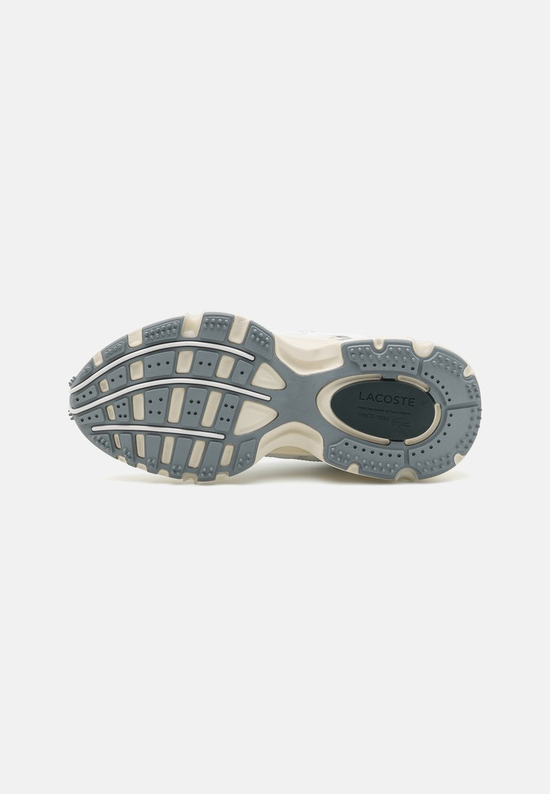 Semelle de chaussure avec une base en caoutchouc gris, présentant un motif de semelle pour une meilleure adhérence, des accents en couleur crème et un logo de marque sur le talon.