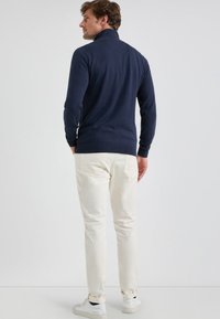 Man in een marineblauwe coltrui, met off-white broek en witte sneakers, staand met zijn handen in zijn zakken, zijwaarts gekeerd.