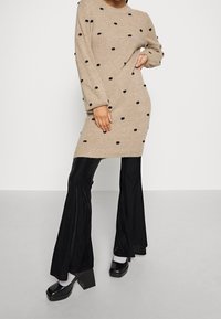 Robe en maille beige à pois noirs, manches longues, coupe décontractée, associée à un pantalon noir évasé et des chaussures à talons épais.