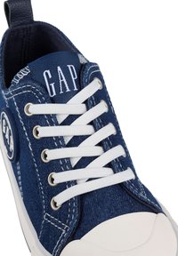 Denim sneaker with white laces, round toe, rubber sole, and blue fabric upper. Features logo on tongue and side. Stitched accents are visible.