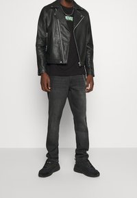 Černá kožená motorkářská bunda, černé grafické tričko, tmavě šedé džíny a černé tenisky. Mezi rysy patří zipy a řetězové detaily.