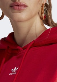 Červená bavlněná mikina s kapucí se stahovací šňůrkou na krku a bílým logem adidas. Zlatá kruhová náušnice zvýrazňuje hladkou texturu pokožky.