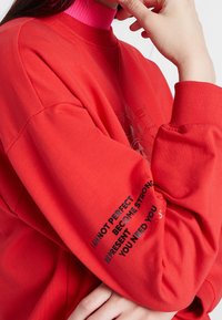 Rode sweatshirt met een gestructureerd oppervlak, geribbelde manchetten en een roze hoge kraag. Heeft bedrukt zwart tekst op de linker mouw.