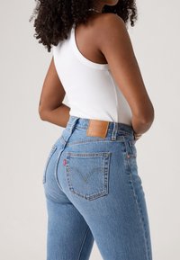 Jeans in denim blu a vita alta con un lavaggio chiaro, caratterizzati da una toppa in pelle, tasche posteriori e una silhouette aderente. Canotta bianca a coste visibile.