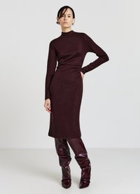 Abito lungo a maniche lunghe color bordeaux in maglia, con collo alto, silhouette aderente e lunghezza midi. Abbinato a stivali lucidi al ginocchio dello stesso colore.