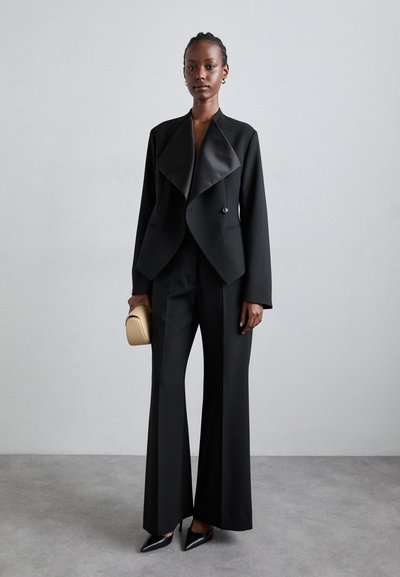 Μαύρο σακάκι προσαρμοσμένο με κολάρο σε ρίγες, συνδυασμένο με παντελόνι σε φαρδιά γραμμή. Το μοντέλο κρατάει μια μικρή μπεζ τσάντα, συμπληρωμένη με μαύρες γόβες με αιχμηρή μύτη.