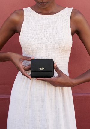 Person i hvid ærmeløs kjole holder en sort pung med gyldent logo mod en rød væg.