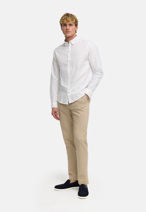 Blondeman staat met handen in de zakken, draagt een wit overhemd met knopen, beige broek en zwarte instapschoenen tegen een effen achtergrond.