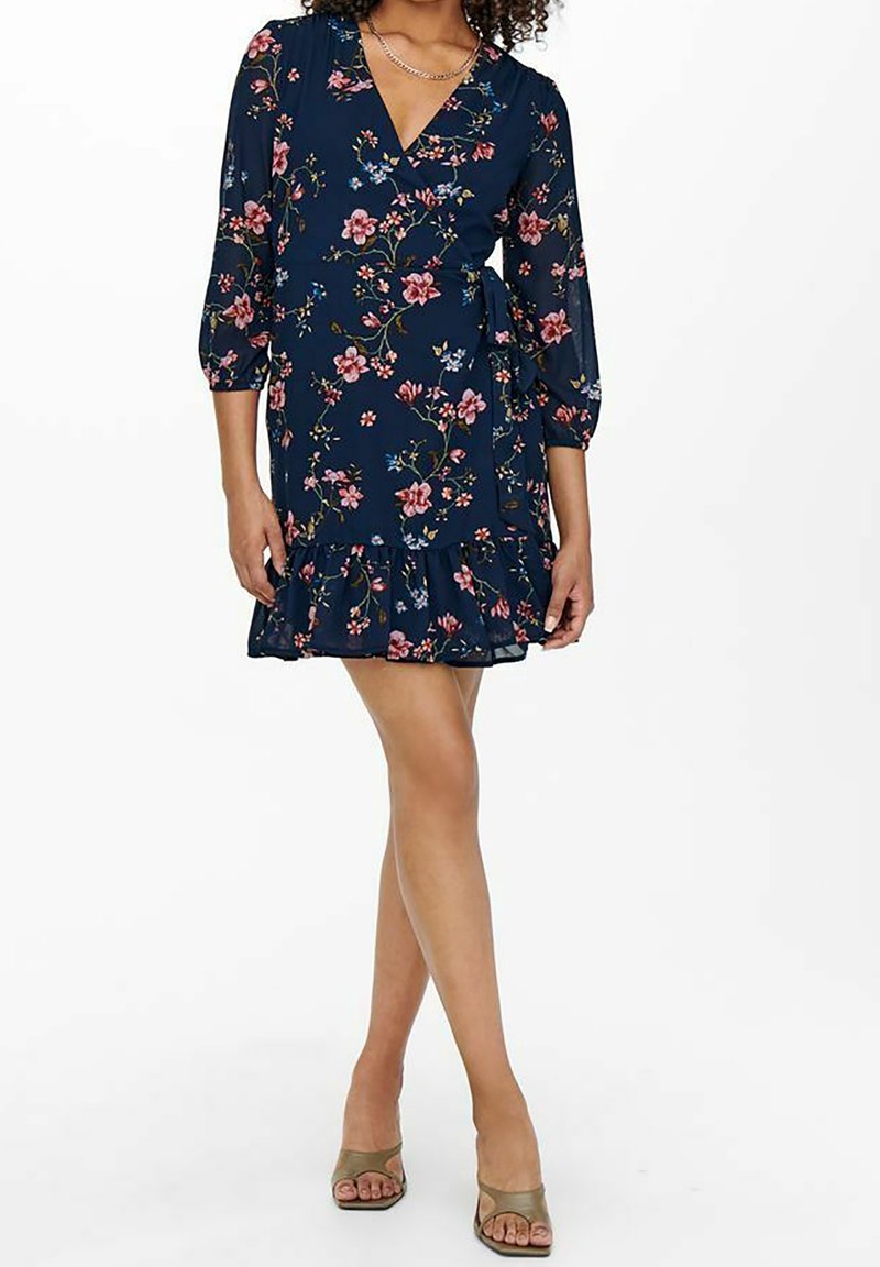 Femme portant une robe portefeuille bleu marine à imprimé floral avec manches trois-quarts et ourlet volanté, associée à des talons ouverts beiges.