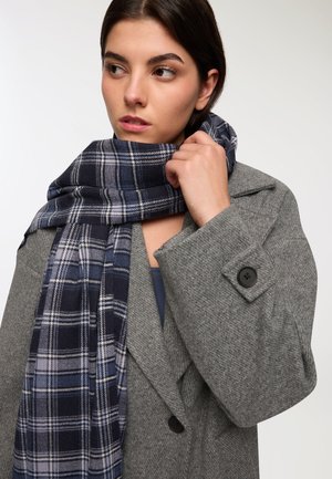 Cappotto di lana grigio con bottoni neri, abbinato a una sciarpa in tartan blu navy con accenti bianchi e azzurri chiari, drappeggiata attorno al collo.