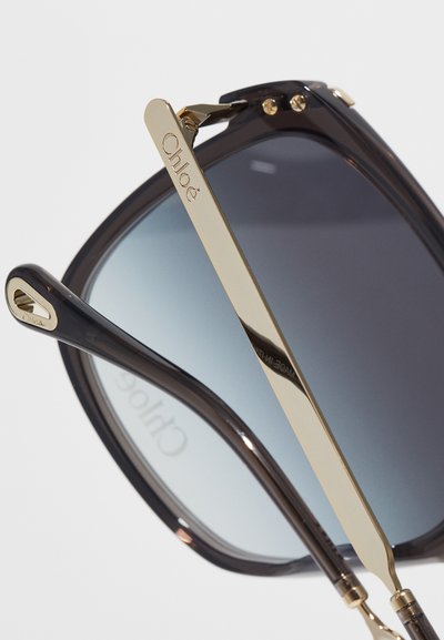 Chloé-solglasögon har en mörk acetatram med gradientblå linser, guldtonade metallarmar och subtil logotypdetaljering på skalmarna.