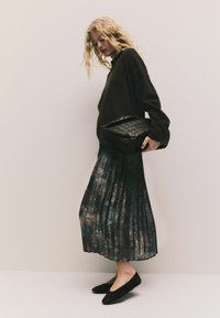 Schwarzer übergroßer Pullover, floral geblümter, plissierter Midi-Rock in Dunkelgrün, schwarze geflochtene Clutch-Tasche und schwarze Ballerinas. Strukturierte Oberflächen mit einem sanften Aussehen.