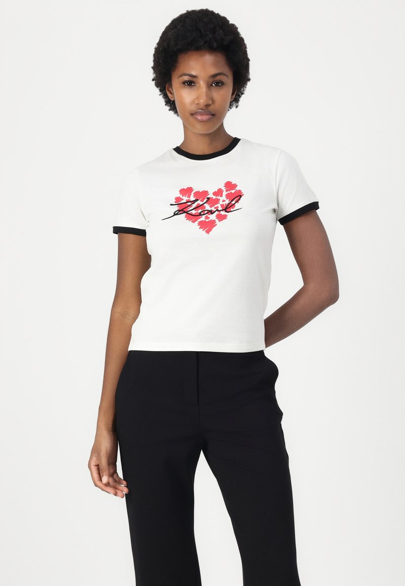 Weißes T-Shirt mit kurzen Ärmeln und schwarzer Einfassung, das ein rotes Herzdesign und den Schriftzug "Karl" zeigt. Aus Baumwolle, taillierte Passform.