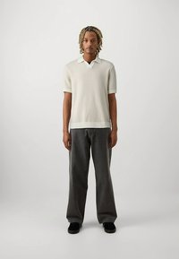 Short-sleeve, light beige knit polo with a ribbed hem, paired with loose-fitting, dark gray jeans and black shoes.