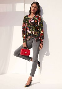 Schwarz mit floralem Muster gestrickter Pullover, graue Jeans, schwarze High Heels mit goldenen Spitzen und eine rote gesteppte Handtasche. Das Model lehnt an einer weißen Wand.