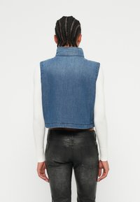 Denim cropped vest med krage, med en blå teksturert finish, kombinert med slanke svarte skinnbukser, sett fra baksiden.