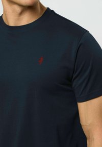 Vīrietis valkā tumši zilu t-kreklu ar apaļu kaklu, ar mazu sarkanu izšūtu logotipu kreisajā krūtīs un īsām piedurknēm.