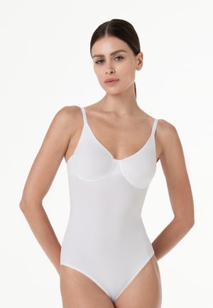 Donna con capelli lisci e scuri che indossa un body bianco aderente con spalline sottili, in piedi davanti a uno sfondo bianco semplice.