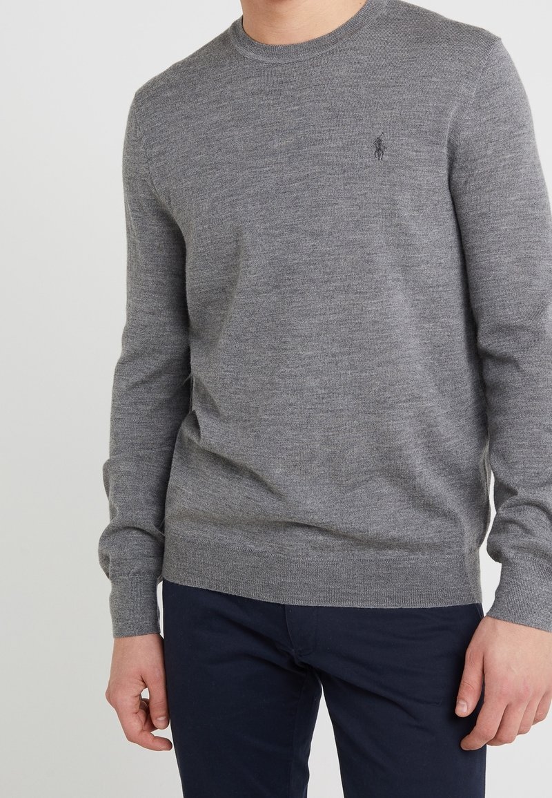 Homme portant un pull gris ajusté à manches longues avec col rond et un pantalon bleu foncé, debout les bras détendus le long du corps.