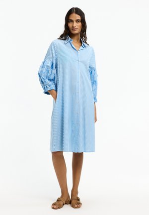 Frau trägt ein blau-weiß vertikal gestreiftes Hemdkleid mit puffärmeligen 3/4-Ärmeln und beigen flachen Sandalen, steht mit einer Hand in der Tasche.