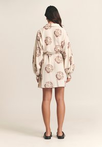 Kreminės spalvos ilgarankovė suknelė, puošta rudos spalvos gėlių raštu, su juosmens diržu, laisvu siluetu ir tekstūruota medžiaga. Juodi, lengvai dėvimi bateliai.
