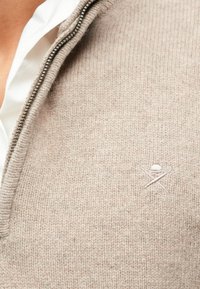 Bežs adīts puloveris ar rāvējslēdzēju, ar teksturētu rakstu un mazu izšūtu logo kreisajā krūtī. Mīksta, ikdienišķa auduma.