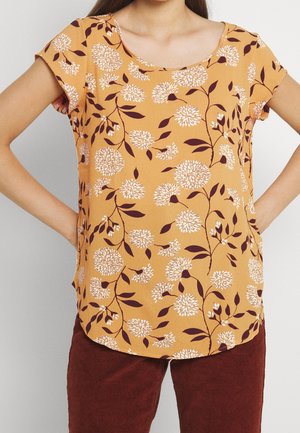 Kvinde iført sennepsgul kortærmet bluse med hvidt blomsterprint og brune manchesterbukser, hænderne hvilende på hofterne.