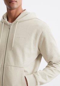 Felpa con zip color beige chiaro realizzata in materiale morbido con una texture liscia, caratterizzata da una tasca frontale e un delicato logo del marchio.